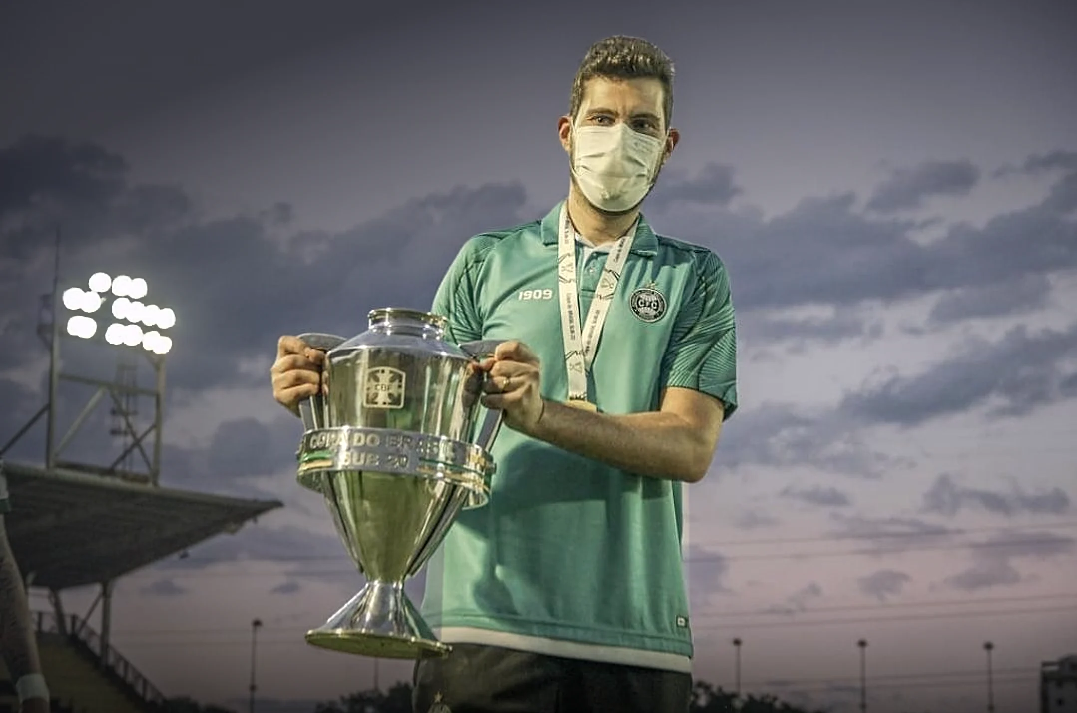 Dr. Marcos Paulo Vanzin de camisa do Coritiba e máscara, segurando uma taça no gramado, com os refletores do estádio acesos ao entardecer